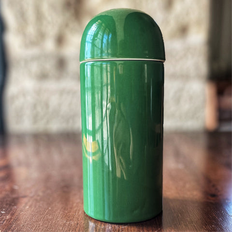 A green, cylindrical Villa Cappelli ceramic olive oil bottle with a glossy finish, crafted by Italian craftsmen, standing on a wooden table.
