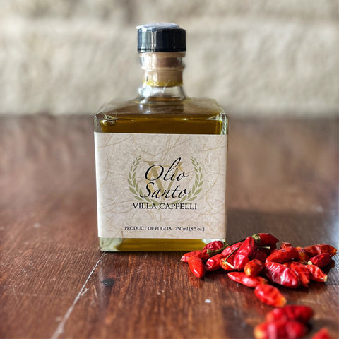 A bottle of Villa Cappelli Olio Santo extra virgin olive oil next to scattered red chili peppers on a wooden surface.