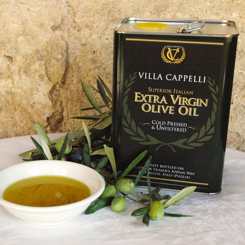 A 3L Tin of Villa Capelli Extra Virgin Olive Oil beside a bowl of olive oil and fresh DOP Italian olives, with an olive branch on a stone surface.