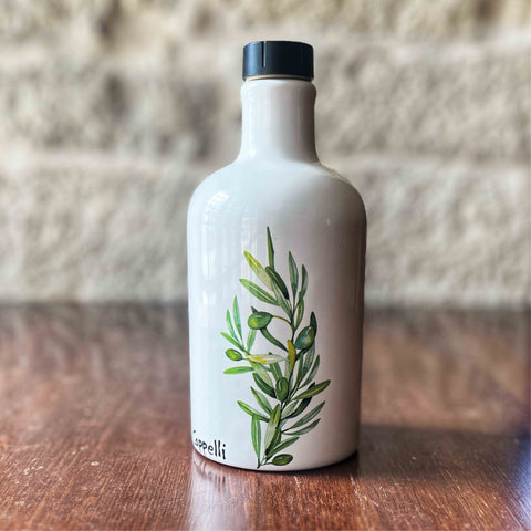 White ceramic olive oil bottle with a green olive branch design displayed on a wooden surface against a brick wall background by Villa Cappelli.