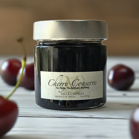 A jar of Villa Cappelli cherry conserves on a wooden table, surrounded by fresh cherries, with a label stating "no preservatives, no artificial ingredients.