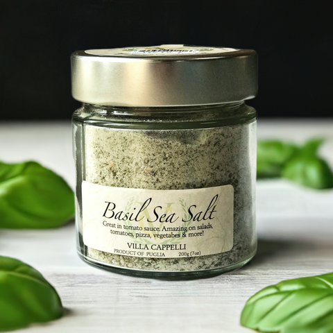 A glass jar of Villa Cappelli Basil Sea Salt with fresh basil leaves around it, labeled for enhancing tomato sauce, pizza, and vegetables.
