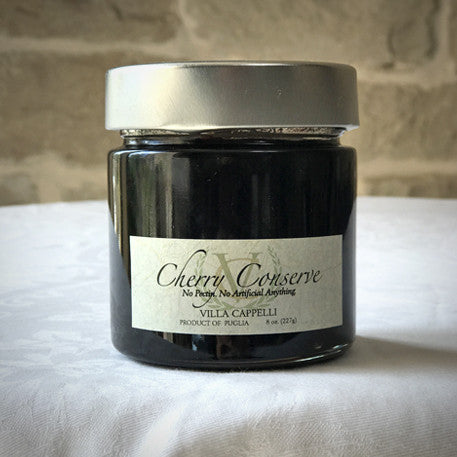 A jar of Villa Cappelli Cherry Conserves, product of Puglia, 8 oz with a gray lid, placed against a stone wall background.