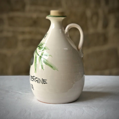 White ceramic Villa Cappelli olive oil jug with a green leaf design and the word "nerine" hand-painted on its surface, displayed on a white cloth against a brick wall background.