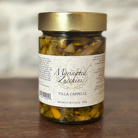 A jar of Villa Cappelli Marinated Zucchini, featuring extra virgin olive oil, displaying a label with product details, set against a wooden backdrop.