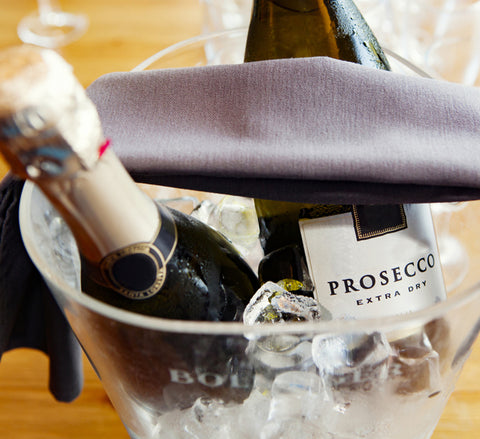 A bucket filled with ice, containing a bottle of Villa Cappelli Prosecco Vinegar and a bottle of champagne, with a cloth napkin on top.