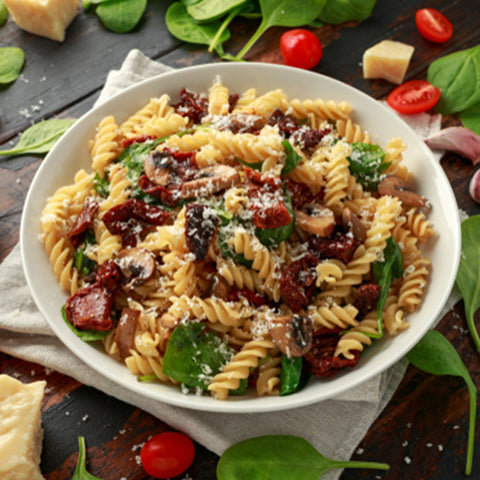 A bowl of pasta with spinach, mushrooms and parmesan cheese mixed with Villa Cappelli Sun-Dried Tomatoes on a wooden table.
