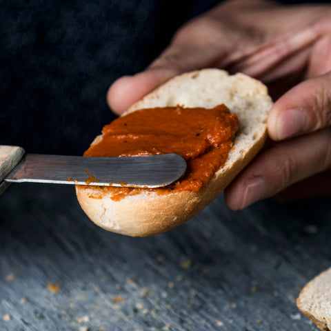 A person cutting a piece of bread with a knife, spreading Villa Cappelli's Spicy Sun-Dried Tomato Spread on it.