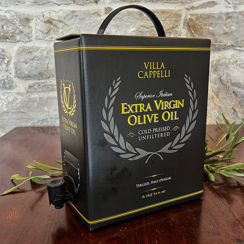A 3L box of Villa Cappelli Extra Virgin Olive Oil with a spout rests on a wooden table, set against a stone wall backdrop.
