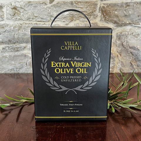 A 3L Villa Cappelli Extra Virgin Olive Oil box, black with gold and white text, sits on wood with olive branches nearby and a stone wall behind—cold pressed Italian oil in an easy-pour package, perfect for your kitchen.