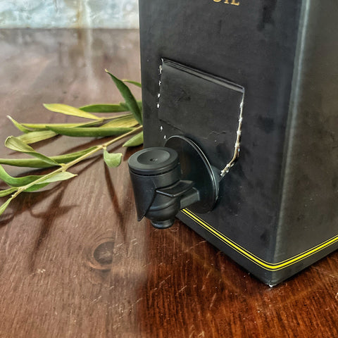 A close-up of a 3L Villa Cappelli Extra Virgin Olive Oil box with a plastic spout dispenser on a wooden surface, highlighted by green leaves beside it.