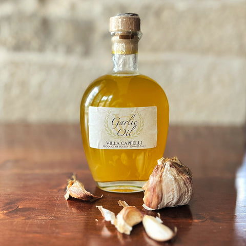 A bottle of Villa Cappelli Garlic Oil sits on a wooden surface next to a whole garlic bulb and cloves, bringing aromatic flavor to any kitchen.