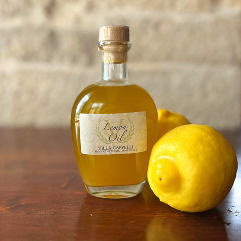 A bottle of Villa Cappelli Lemon Oil rests on a wooden surface next to two whole lemons, set against a neutral background, showcasing its fresh flavor.