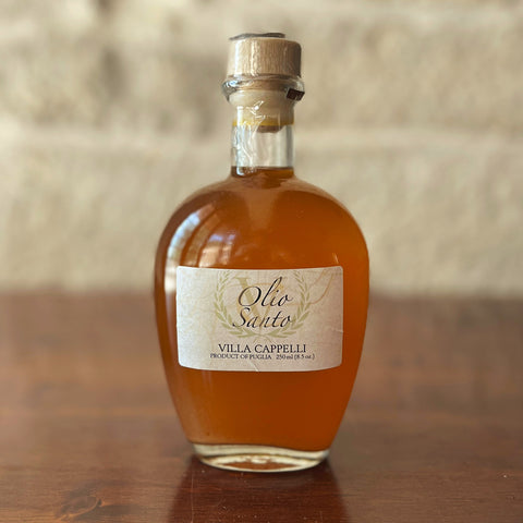 A clear glass bottle of Villa Cappelli Olio Santo, an extra virgin olive oil infused with chili peppers and topped with a cork stopper, rests on a wooden surface against a neutral background.
