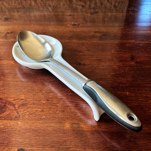A Villa Cappelli Pausa Perfetta metal spoon with a black handle rests on an artisanal spoon rest, handcrafted in Puglia, atop a wooden table.