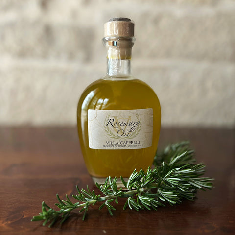 A glass bottle of Villa Cappelli Rosemary Oil sits on a wooden surface, with a sprig of fresh rosemary in front.