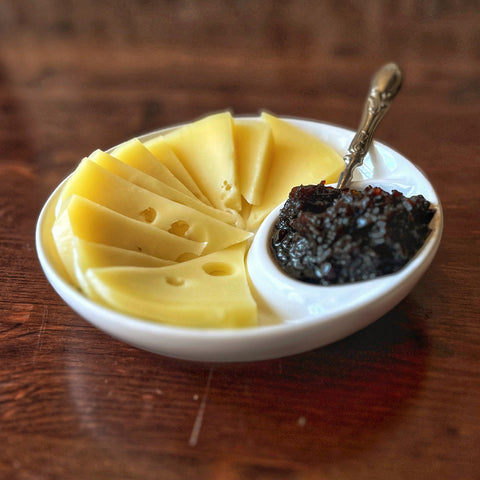 The Villa Cappelli Spuntino Server, a handcrafted white ceramic piece, displays Swiss cheese on one side and dark chutney or jam on the other, arranged atop a wooden surface.