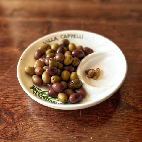 The Villa Cappelli Spuntino Server, a round handcrafted ceramic plate holding assorted green and purple olives with olive pits on the side, adds Mediterranean charm to your table.