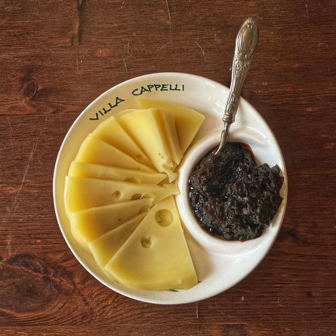The Villa Cappelli Spuntino Server, a handcrafted ceramic dish, presents fanned sliced cheese with chutney or jam and a small spoon, bringing Mediterranean charm to the wooden table.