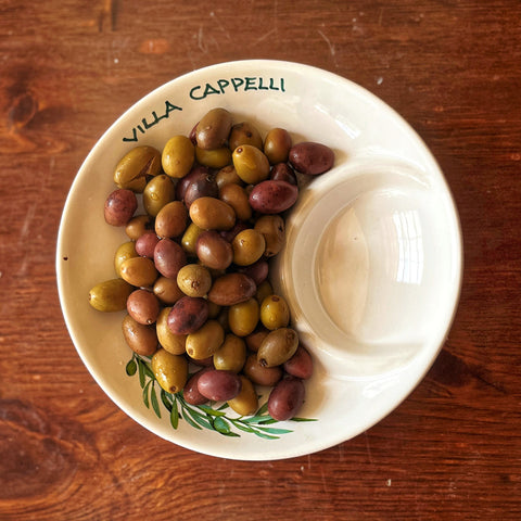 The Villa Cappelli Spuntino Server, a handcrafted ceramic dish, holds green and purple olives on a wooden surface; one empty section of the server enhances its Mediterranean charm.