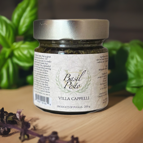 A jar of Villa Cappelli Basil Pesto on a wooden surface with fresh basil leaves and purple flowers in the background.