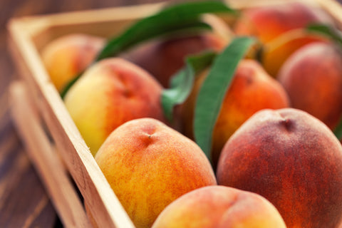 A crate of fresh peaches from Villa Cappelli, some adorned with green leaves, ideal for making their renowned Peach Conserve.